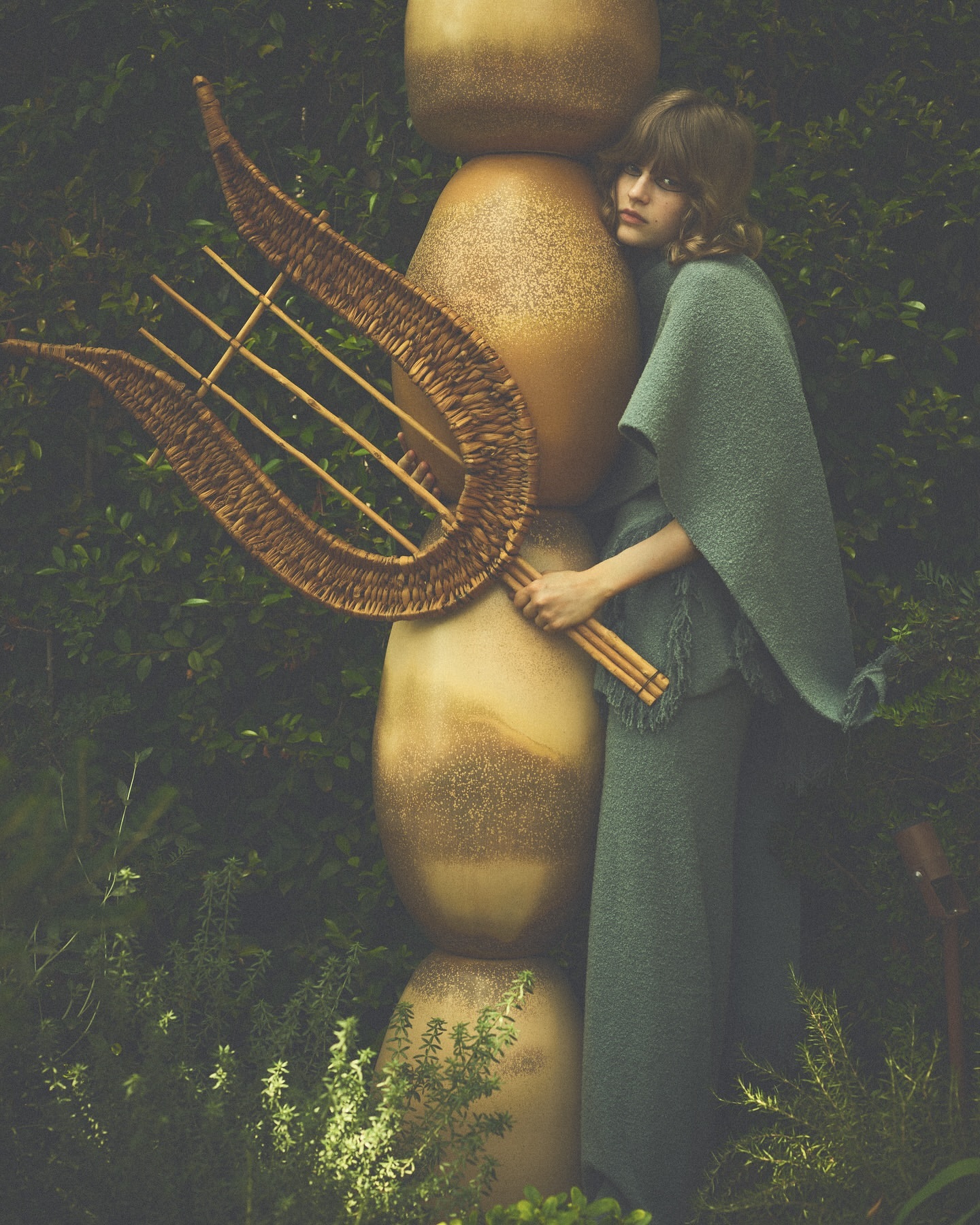 Young woman in green outfit poses with a wicker lyre next to a stack of large ceramic vessels in an overgrown garden setting.