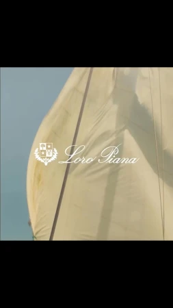 Close-up of a white sail on a boat with Loro Piana logo. The sail is angled, and the background is light blue sky.