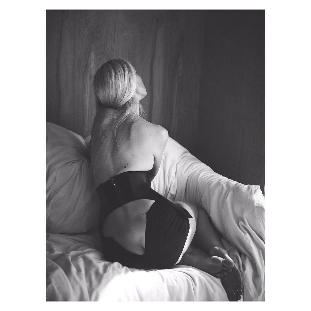 Back view of a blonde woman in black shapewear sitting on a white bed with white curtains in a minimalist, black and white photo.
