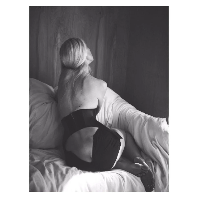 Back view of a blonde woman in black shapewear sitting on a white bed with white curtains in a minimalist, black and white photo.