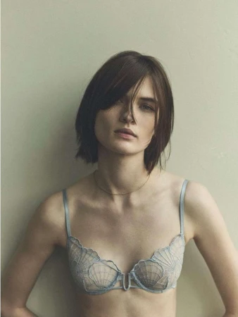 Portrait of a woman with short dark hair wearing a light blue lace bra against a neutral studio background
