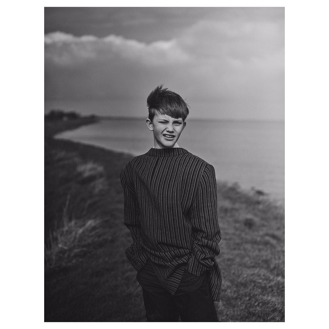 Monochrome portrait of a teenage boy with dark hair in a striped shirt standing outdoors near a body of water under a cloudy sky.