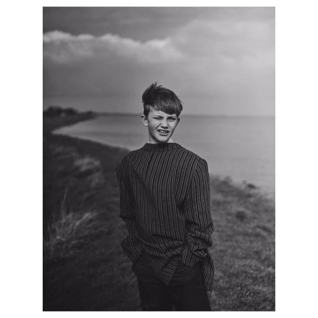 Monochrome portrait of a teenage boy with dark hair in a striped shirt standing outdoors near a body of water under a cloudy sky.