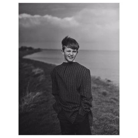 Monochrome portrait of a teenage boy with dark hair in a striped shirt standing outdoors near a body of water under a cloudy sky.