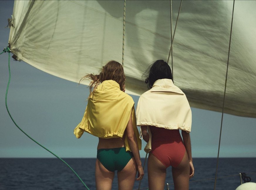 Two women in swimwear and jackets on a sailboat with a large sail against a blue sky, viewed from the back, conveying a sense of travel.