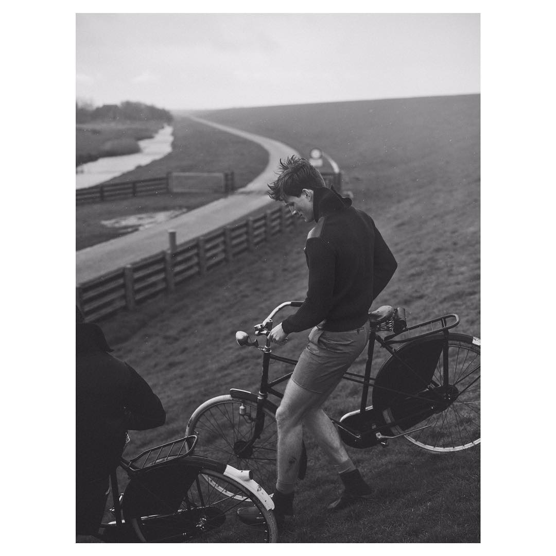 A man walks with his bicycle on a grassy hill in a black and white outdoor shot, with a paved road and overcast sky in the background.