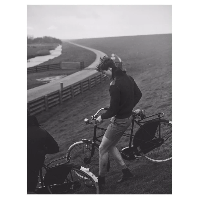 A man walks with his bicycle on a grassy hill in a black and white outdoor shot, with a paved road and overcast sky in the background.