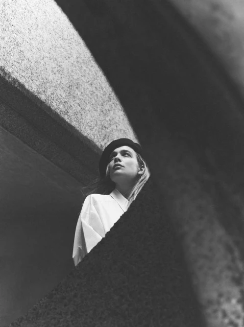 Monochrome portrait of a young adult female in a white shirt and hat, gazing upward in an urban setting with stark, architectural lighting.