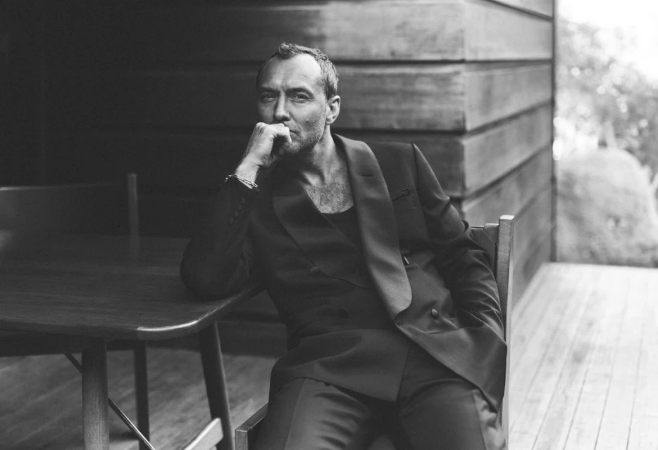 Black and white portrait of a mature adult male wearing a suit, seated at a wooden table in front of a textured wooden wall.
