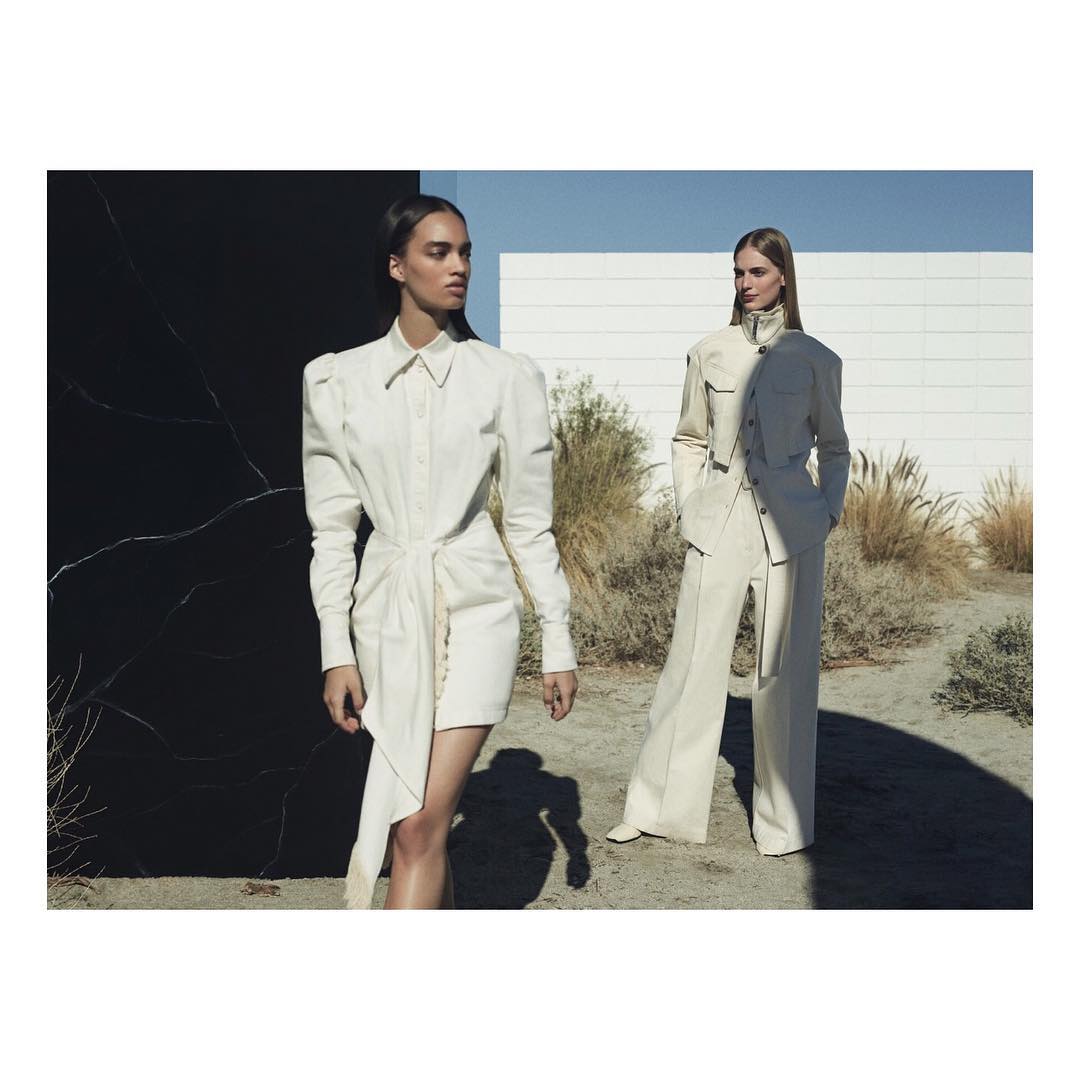 Two fashion models pose outdoors wearing cream-colored outfits against a black marble wall and a white building on a sunny day.