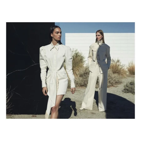 Two fashion models pose outdoors wearing cream-colored outfits against a black marble wall and a white building on a sunny day.