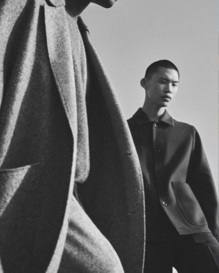 Two Asian male models in a monochromatic fashion shoot featuring wool coats and jackets against a bright, neutral background in a portrait style.
