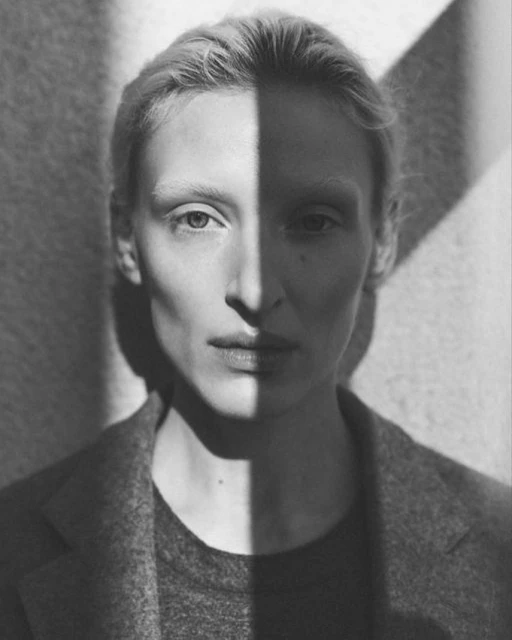 Monochrome portrait of a fair-skinned woman with neutral expression, half of her face lit, wearing a wool coat indoors.