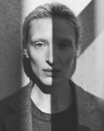 Monochrome portrait of a fair-skinned woman with neutral expression, half of her face lit, wearing a wool coat indoors.