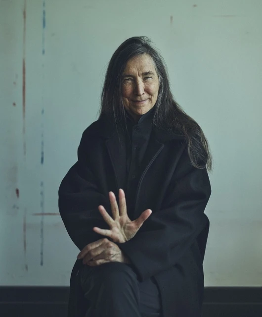 Portrait of an adult woman with long dark hair, wearing a black coat, seated in front of a light-colored wall with paint streaks.