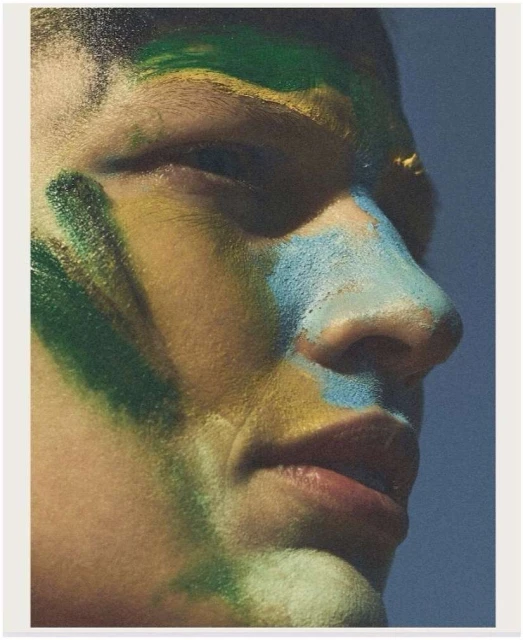 Close-up portrait of an adult male model with vibrant green, blue, and yellow body paint on his face against a blurred light blue backdrop.