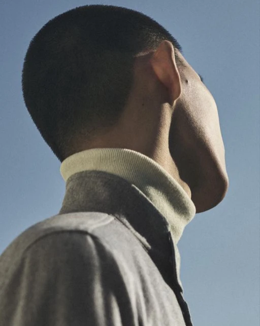 Low-angle shot of an adult man with a buzzcut wearing a collared shirt and jacket against a clear blue sky outdoors. Fashion, style, and menswear.