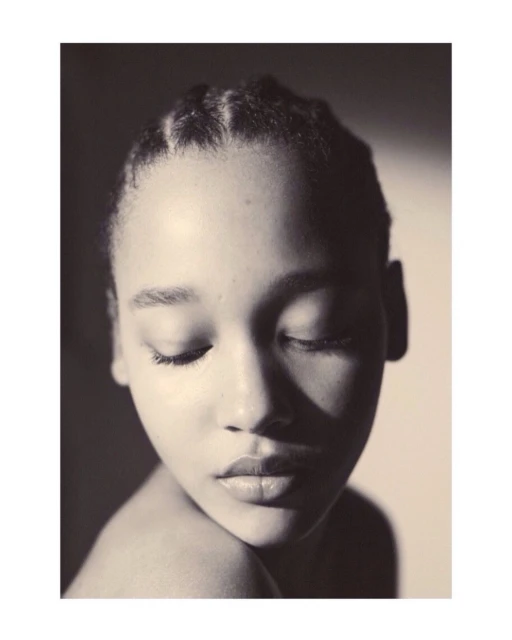 Close-up portrait of a young adult female with dark hair styled in neat braids, eyes closed, and subtle makeup, in a soft, neutral-toned setting.