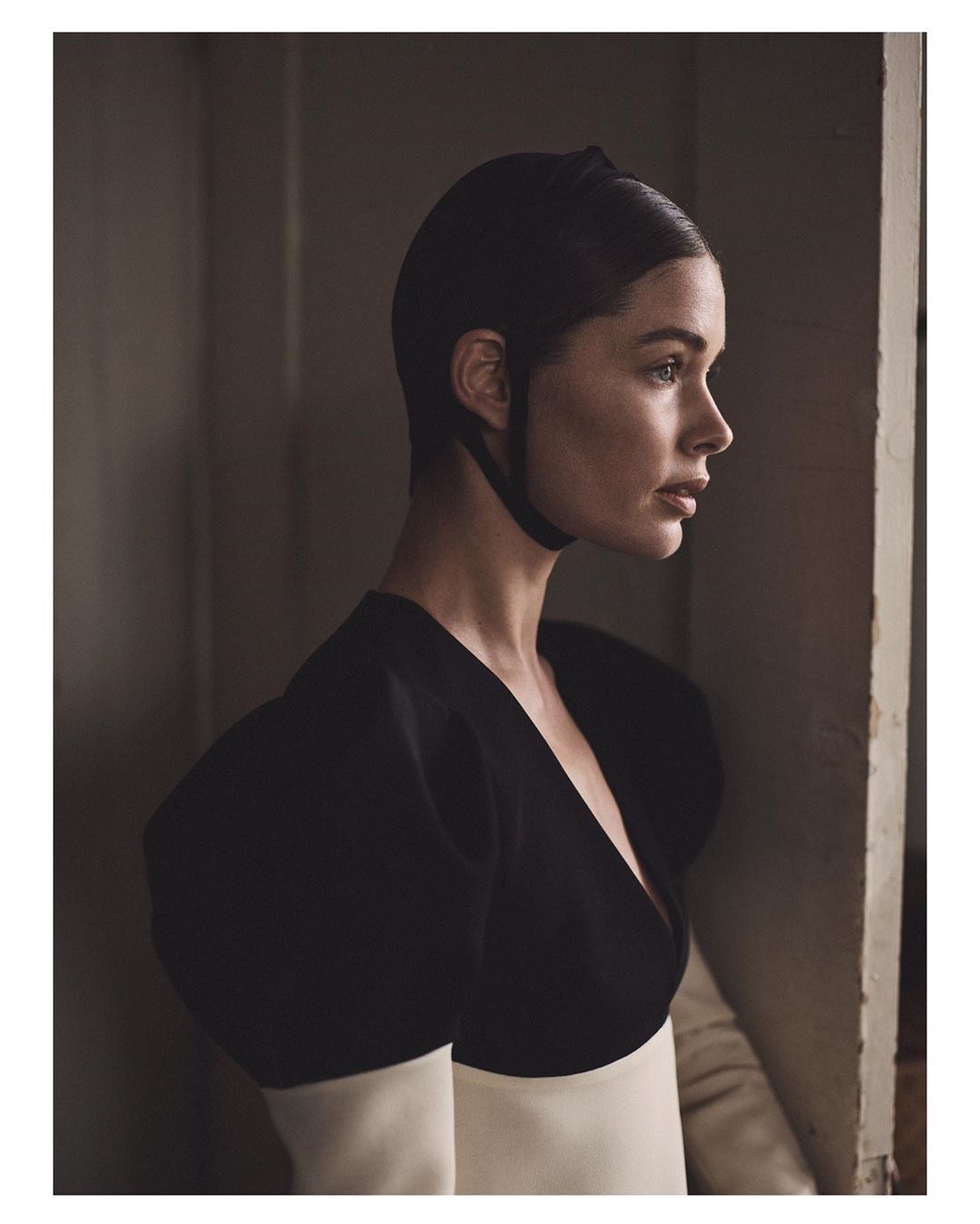 Portrait of a woman with short dark hair wearing a black and cream dress, posing in front of a dark gray wall and looking out a window.