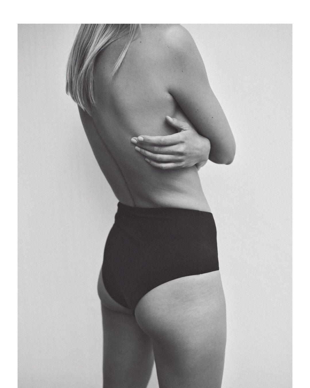 Back view of a fair-skinned adult female wearing black high-waisted bottoms, facing away from the camera with arms crossed.
