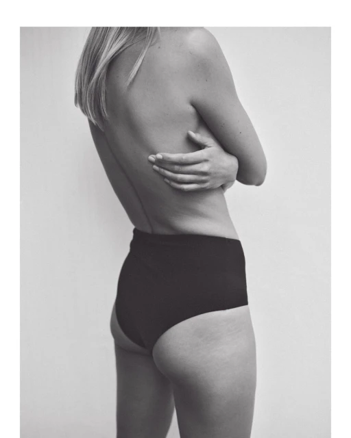 Back view of a fair-skinned adult female wearing black high-waisted bottoms, facing away from the camera with arms crossed.