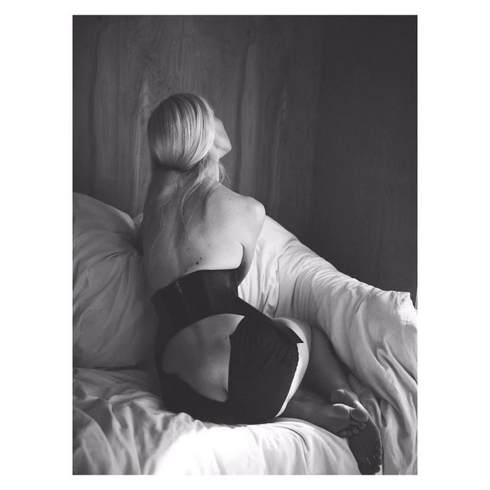 Blonde woman wearing a black corset seated on a plush white bed by Annemarieke Van Drimmelen
