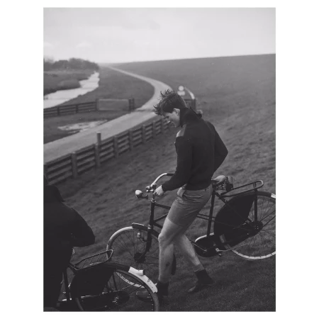 A man standing next to a bicycle on a sloping embankment with a winding road and waterway in the background.