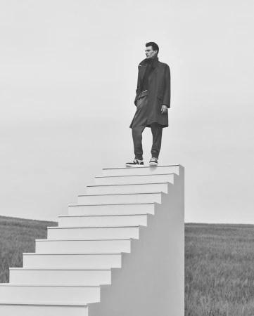 Monochrome portrait of a man standing at the top of a large outdoor white staircase under an overcast sky.