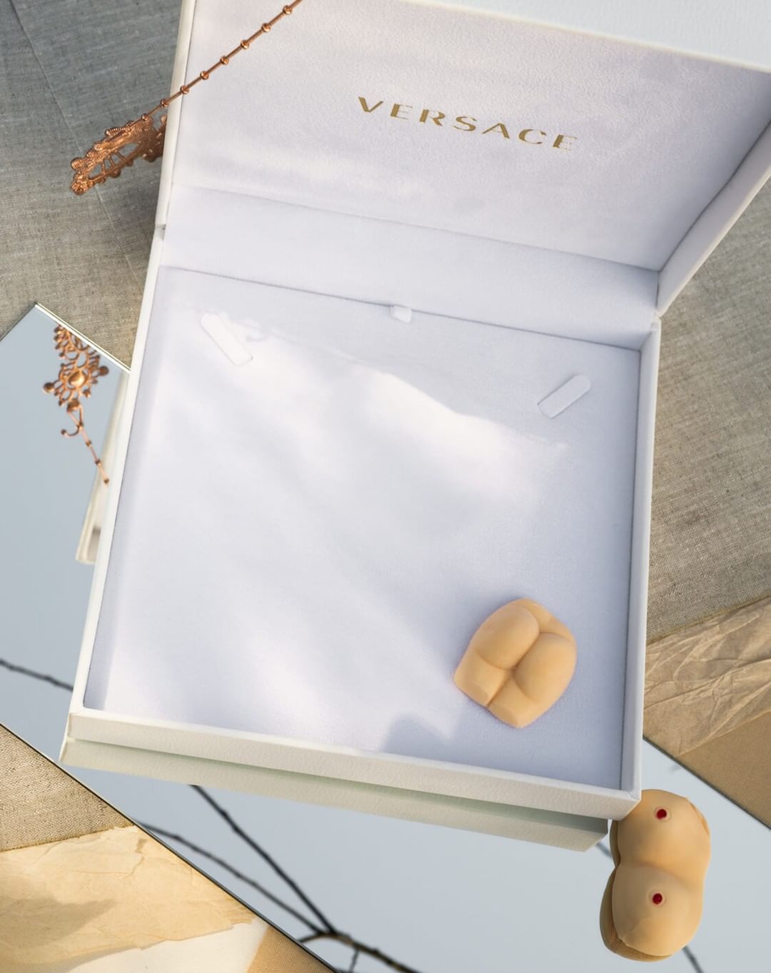 White Versace box with small ceramic butt art object. Neutral beige, white, and grey tones. Artful product display, indoor setting, studio lighting.