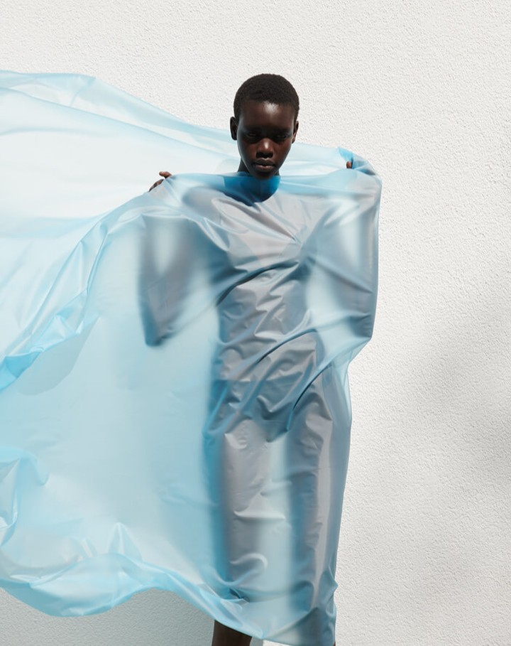 Adult female with dark skin and short hair stands in front of a white wall, covered in a sheer, light-blue, flowing fabric dress.