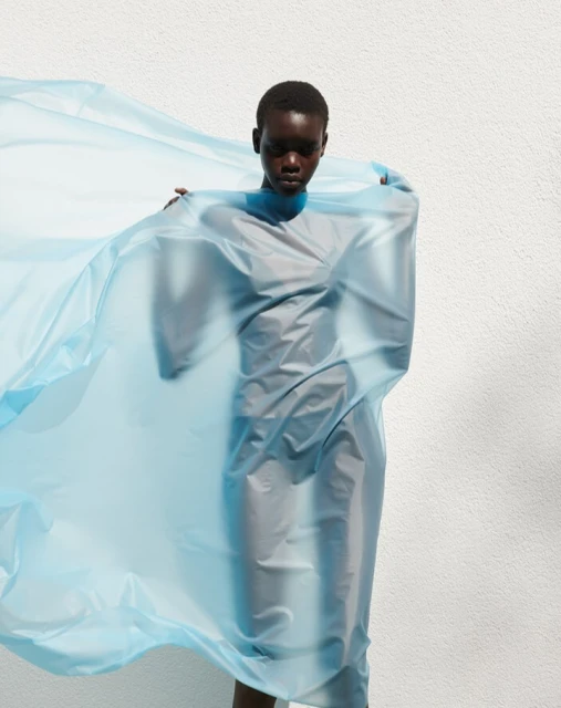 Adult female with dark skin and short hair stands in front of a white wall, covered in a sheer, light-blue, flowing fabric dress.