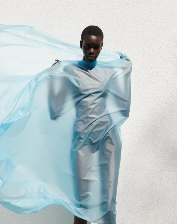 Adult female with dark skin and short hair stands in front of a white wall, covered in a sheer, light-blue, flowing fabric dress.