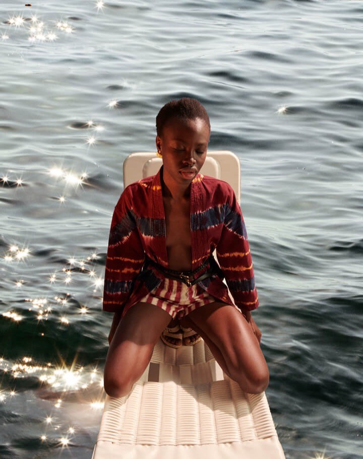 Woman kneeling on a diving board wearing a patterned jacket and shorts, with the sea as a backdrop in sun dappled light.