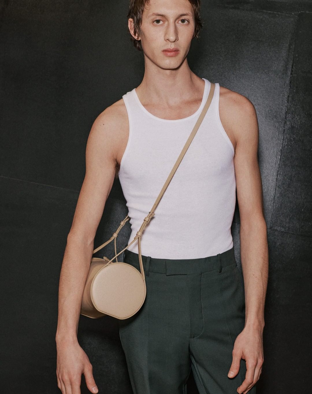 Studio shot of an adult male in a white tank top and dark green pants posing with a beige, cylindrical cross-body bag against a dark background.
