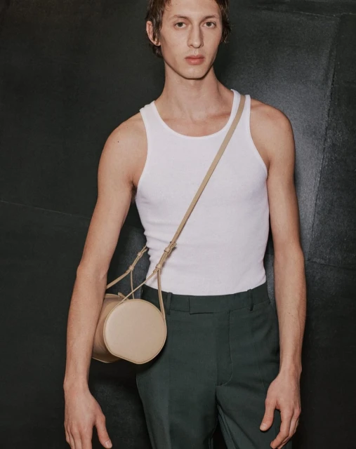 Studio shot of an adult male in a white tank top and dark green pants posing with a beige, cylindrical cross-body bag against a dark background.