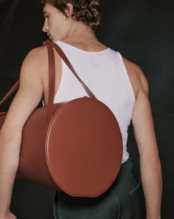 Man wearing a white tank top carrying a large cylindrical brown leather duffel bag over his shoulder against a dark background