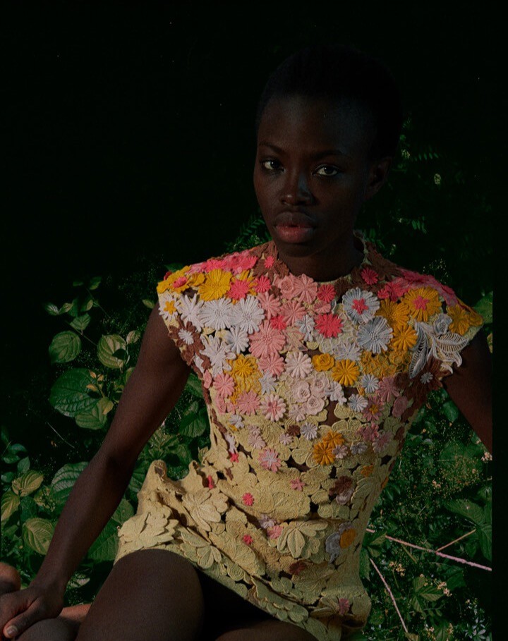 A black model poses in a short yellow dress adorned with colorful flowers against a leafy background.