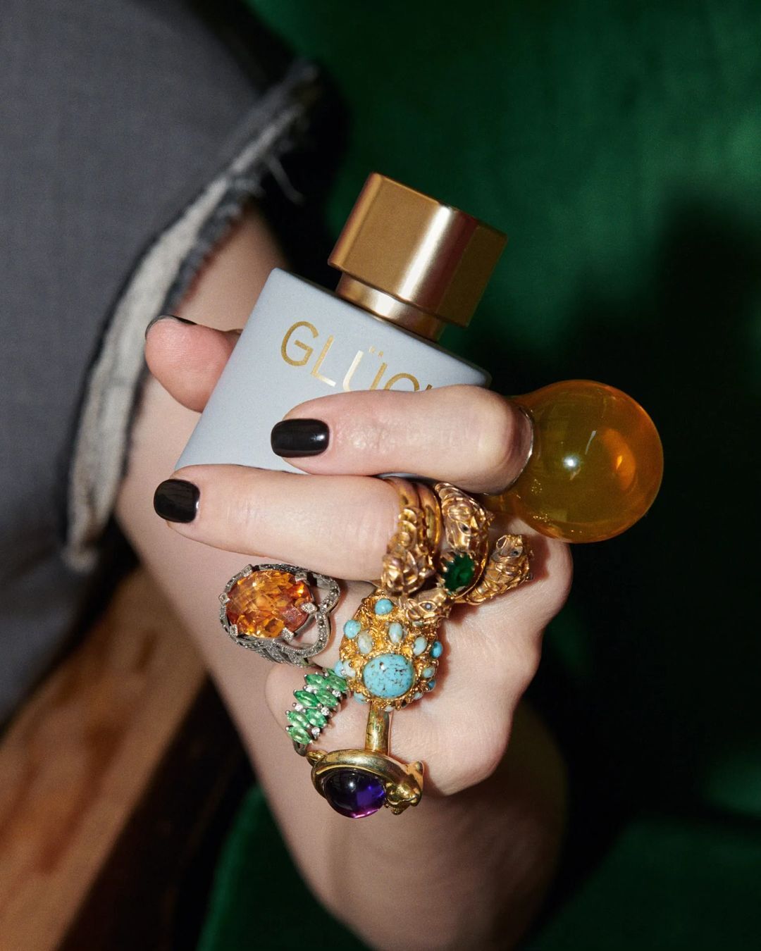 Woman's hand holds grey Gluck perfume bottle with gold cap and decorative rings against blurred green fabric backdrop.