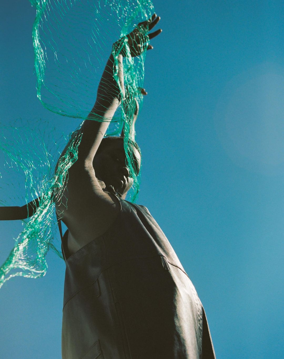 Low angle shot of a Black woman in a gray dress raising her arm up to a turquoise net against a bright blue sky.