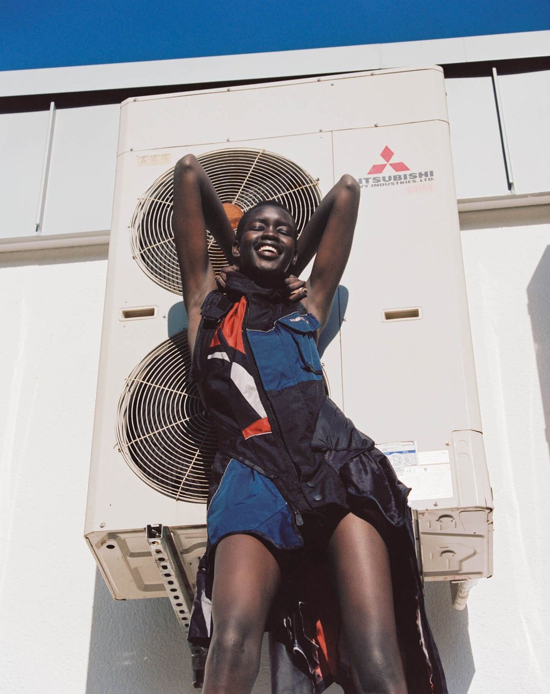 Smiling Black woman in a colorful dress poses outdoors against a white wall and large air conditioning unit under a clear blue sky.