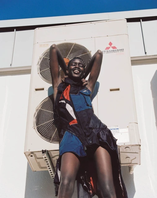 Smiling Black woman in a colorful dress poses outdoors against a white wall and large air conditioning unit under a clear blue sky.
