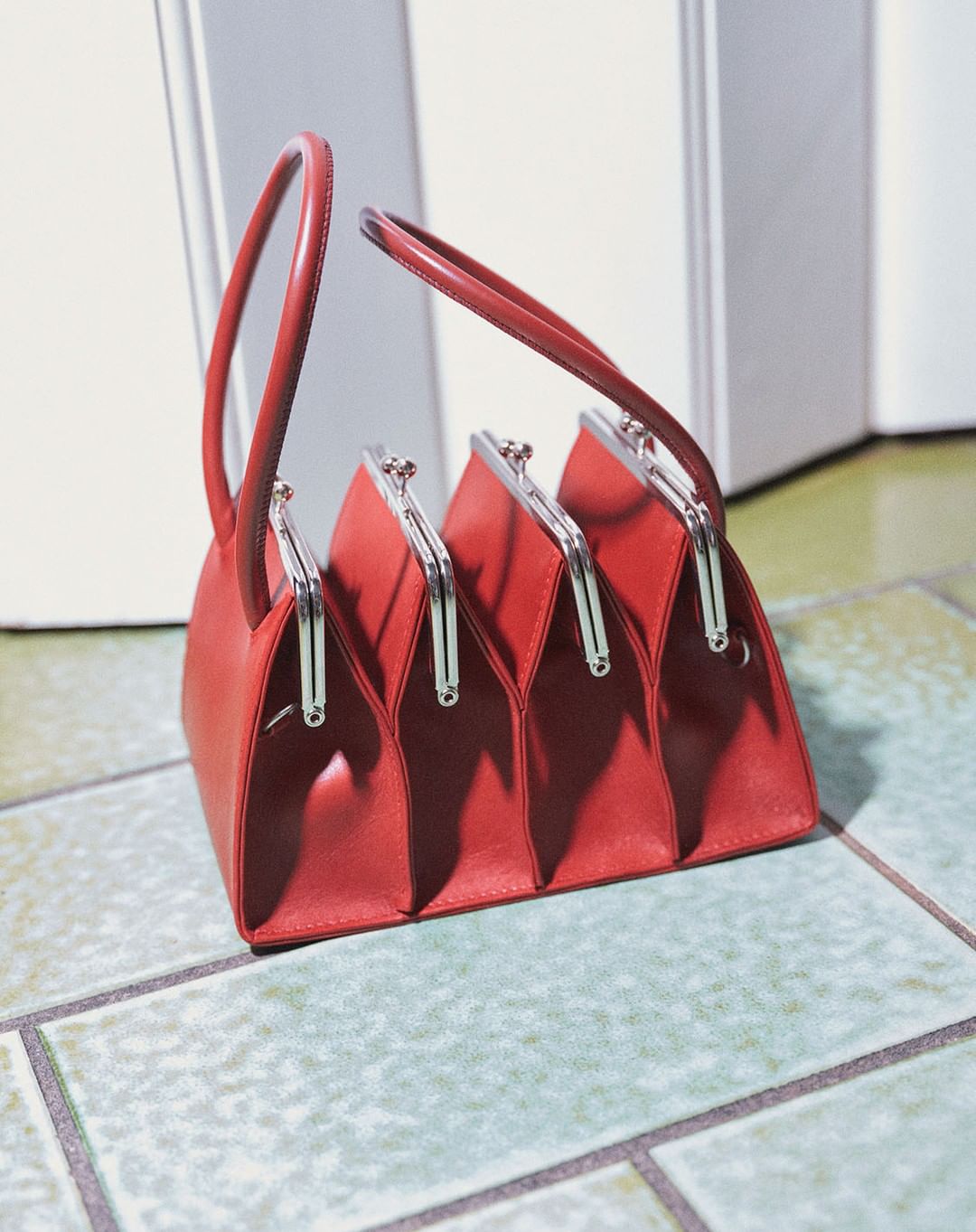 A red leather handbag with four compartments is displayed on a tiled floor against a white wall, showing its unique design and style.