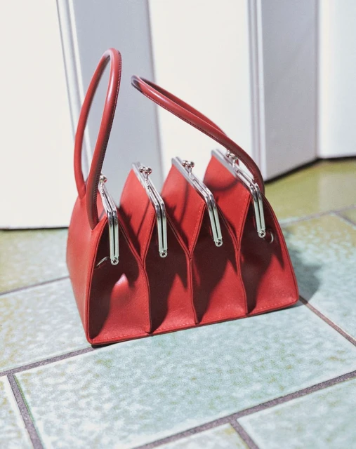 A red leather handbag with four compartments is displayed on a tiled floor against a white wall, showing its unique design and style.