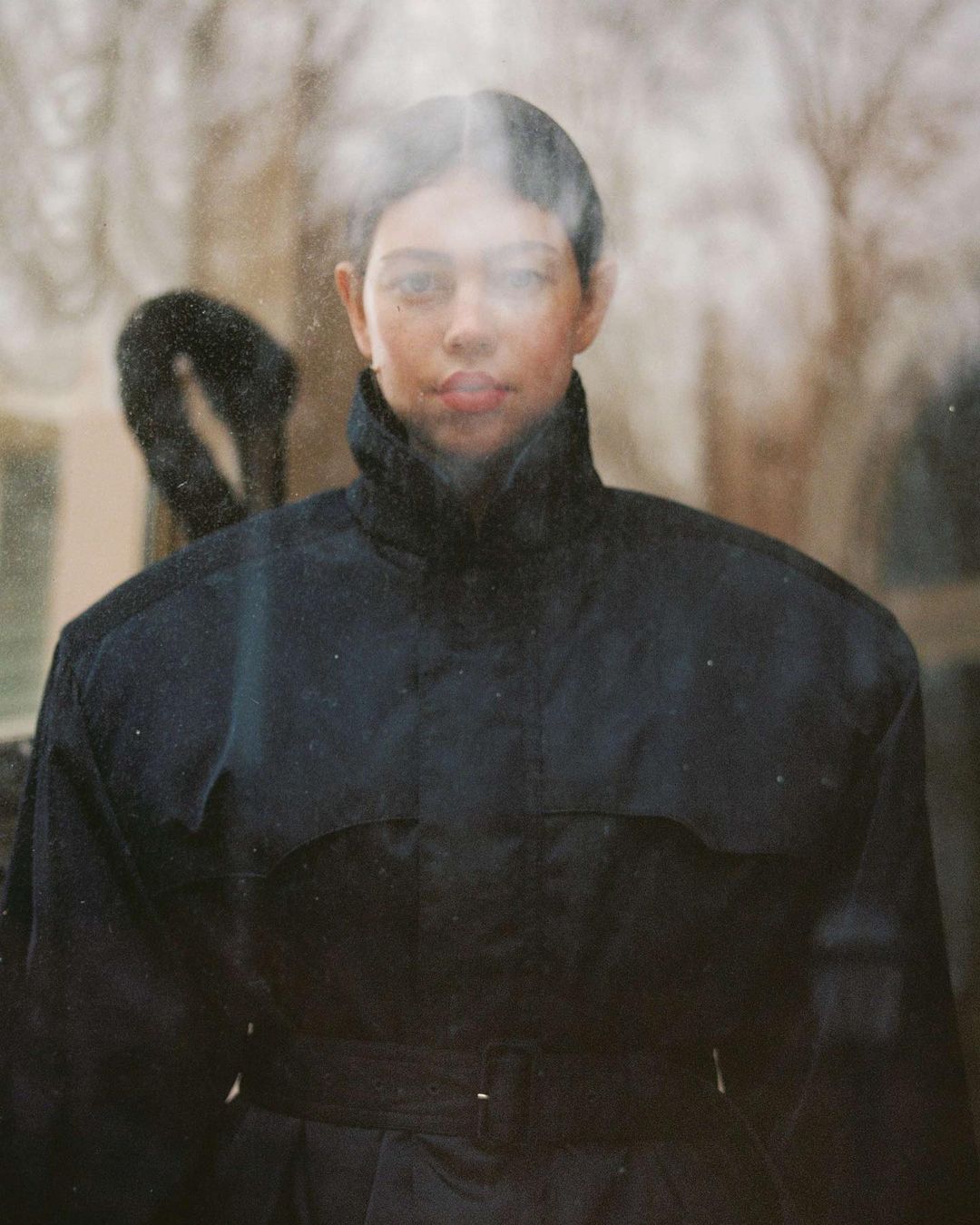 Portrait of a young adult female with short dark hair in an outdoor setting, wearing a dark belted jacket in front of trees.