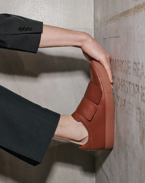 Tan leather slip-on shoe is held against a textured stone wall by a hand clad in a black suit jacket sleeve, footwear fashion.
