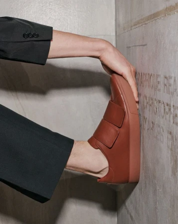 Tan leather slip-on shoe is held against a textured stone wall by a hand clad in a black suit jacket sleeve, footwear fashion.