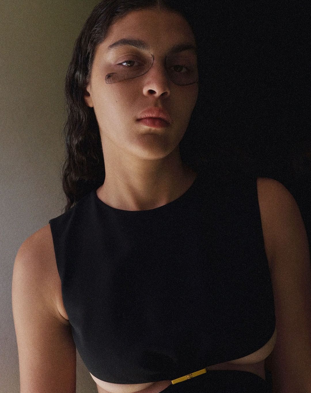 Portrait of a young woman with dark hair and a black sleeveless dress with a cut-out, posing indoors in natural light.