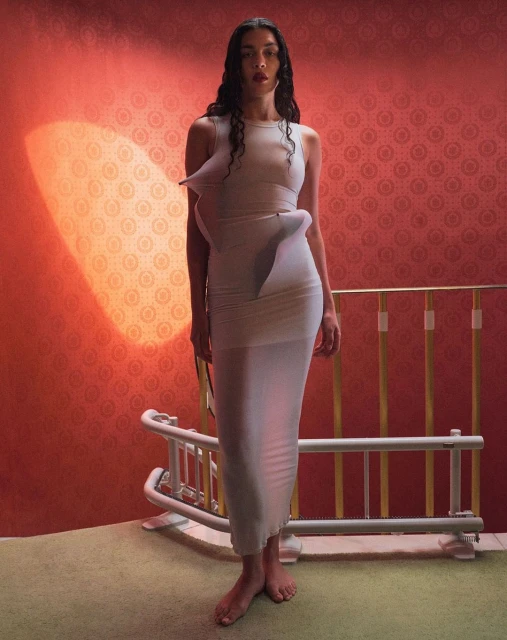 A woman poses in a pale dress in front of an orange wall with a decorative baby cot, creating a stylized and artistic fashion portrait.