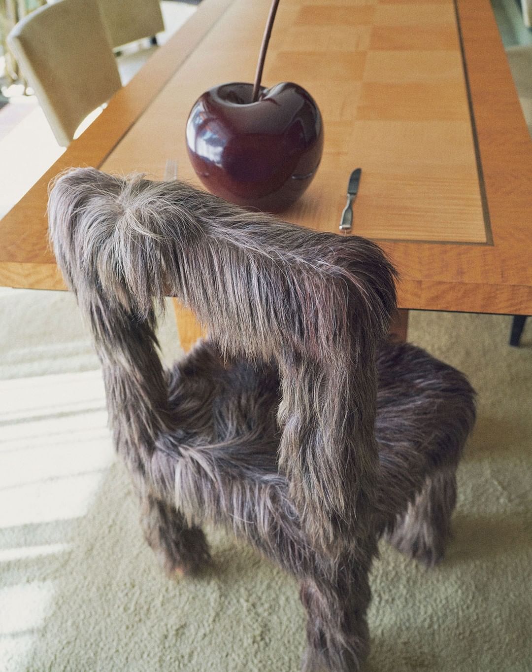 Fur chair with a cherry sculpture on a wood table sits on a carpeted floor in a brightly lit room. Furniture design.
