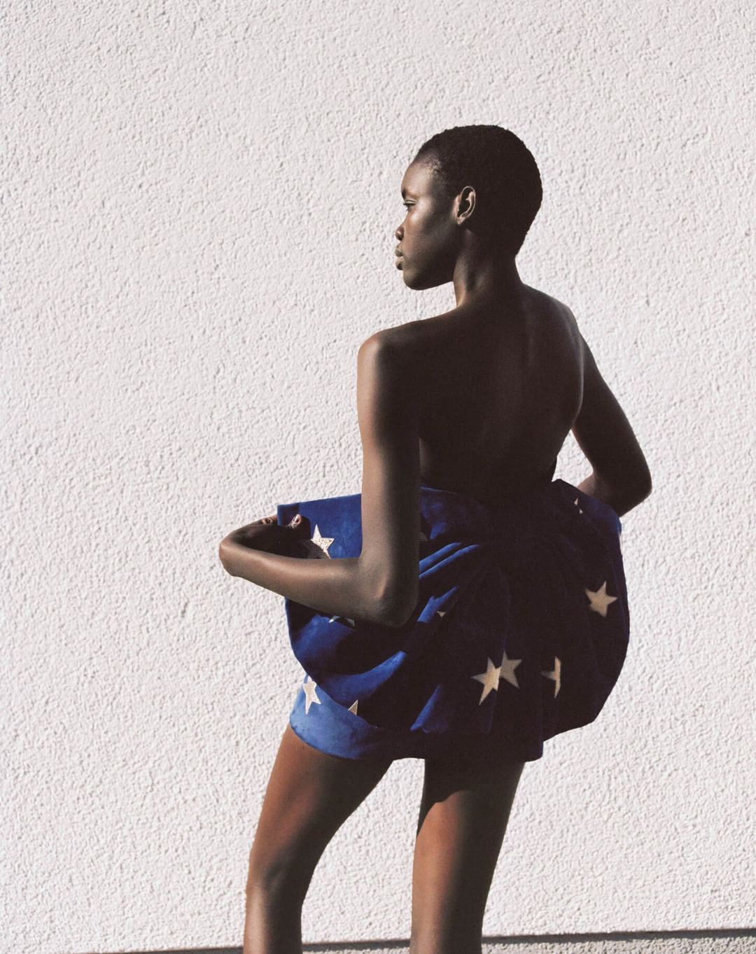 Back view of an African American woman with short hair, draped in blue fabric with white stars against a white wall in natural light.